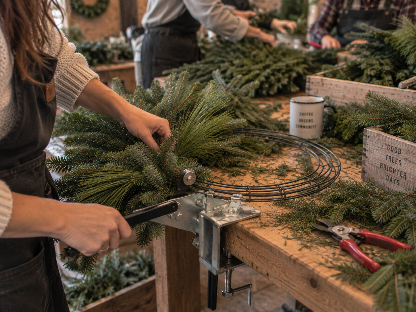 Guests wiring fresh evergreen boughs onto wreath frames at a rustic workshop table